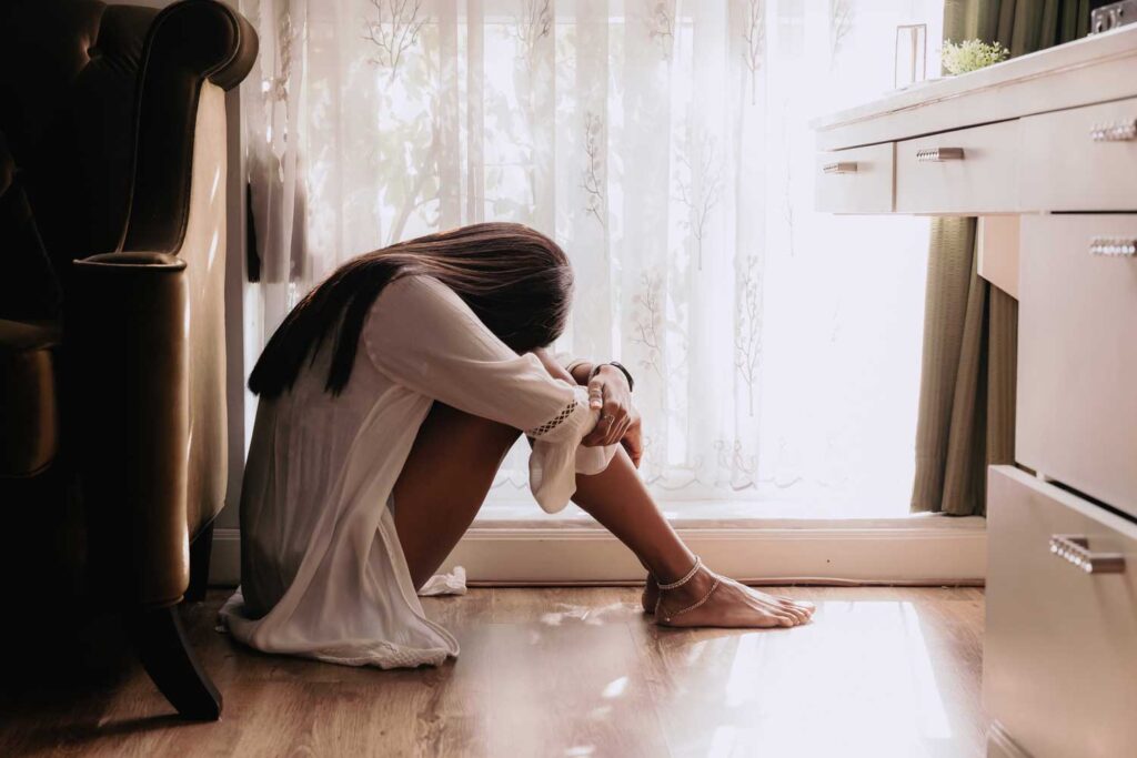 A woman sitting on the floor leaning against a bed, head down, suggesting contemplation or distress in a sunlit room.