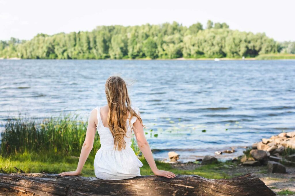 A serene young woman sitting on a log, gazing out at a tranquil lake, reflecting on life's journey amidst nature's calm.